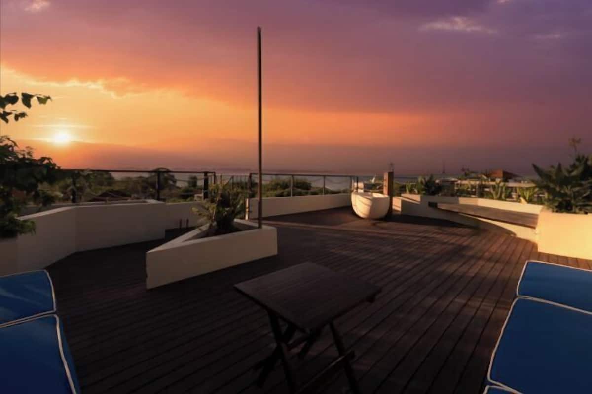 A wooden deck with a table and chairs, surrounded by planters and a railing, overlooking a coastal landscape.