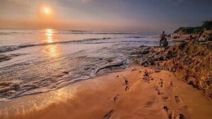 Escena de playa con huellas en la arena, un atardecer y gente al fondo.