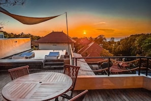 Una terraza en la azotea con mesa y sillas, una hamaca y vistas al atardecer sobre un paisaje con edificios.