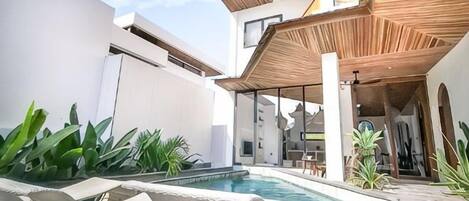 A modern outdoor pool area with lounge chairs, a wooden pergola, and a view of the building's interior.