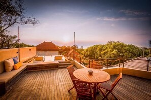 Una terraza de madera con una mesa redonda y sillas, un sofá con cojines, y una barandilla con vistas a un área boscosa.