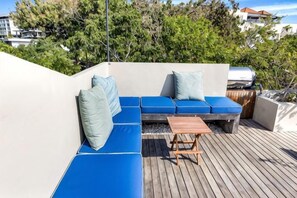 Una terraza de madera con un área de descanso acolchada en tono azul y una pequeña mesa.