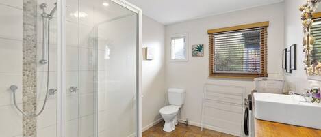 Un baño con ducha envuelta en cristal, inodoro, lavabo y ventana con persianas.