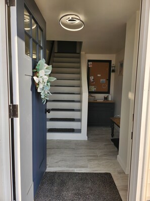 Un escalier menant à un palier où se trouve une porte à gauche, un tableau d’affichage avec des avis, et une petite table avec une lampe.