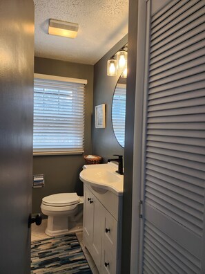 A bathroom with a toilet, sink, and vanity, featuring a window with blinds, a mirror with lights, and a textured ceiling.