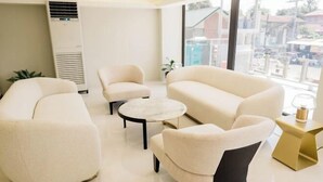A modern hotel room with a large window, a white air conditioner, a sofa, two armchairs, a small round table, and a gold side table with a lamp. - The Manaoag Hotel (Manaoag)