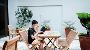 A person sitting at a wooden table with a laptop, surrounded by potted plants and wooden chairs in an outdoor setting. - The Manaoag Hotel (Manaoag)