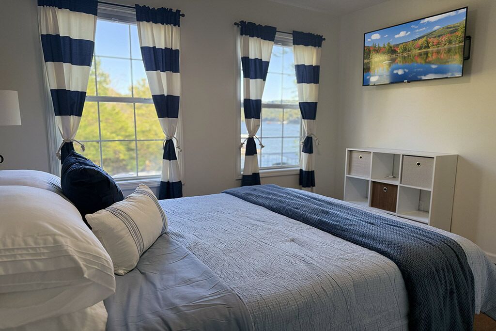 Ein Schlafzimmer mit einem Bett, gestreifte Vorhänge, einem Fernseher an der Wand und Blick ins Freie.