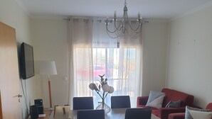 A modern living room with a red sofa, a dining table with chairs, a TV mounted on the wall, and a chandelier. - Wave View Holidays (Albufeira)