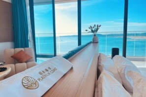 A room with a view of the ocean, a bed with a pillow, a side table, and a vase with flowers.