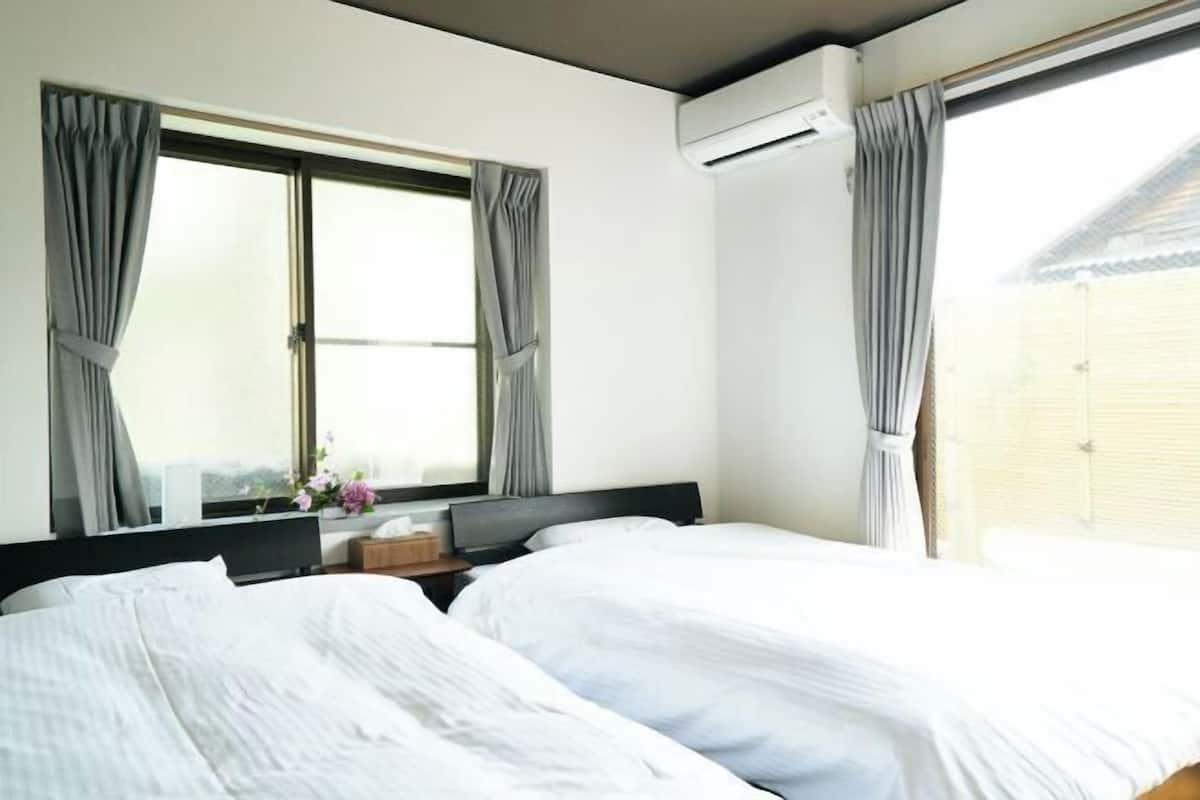 A hotel room with two beds, a window with curtains, an air conditioning unit, and a small table with a vase of flowers.