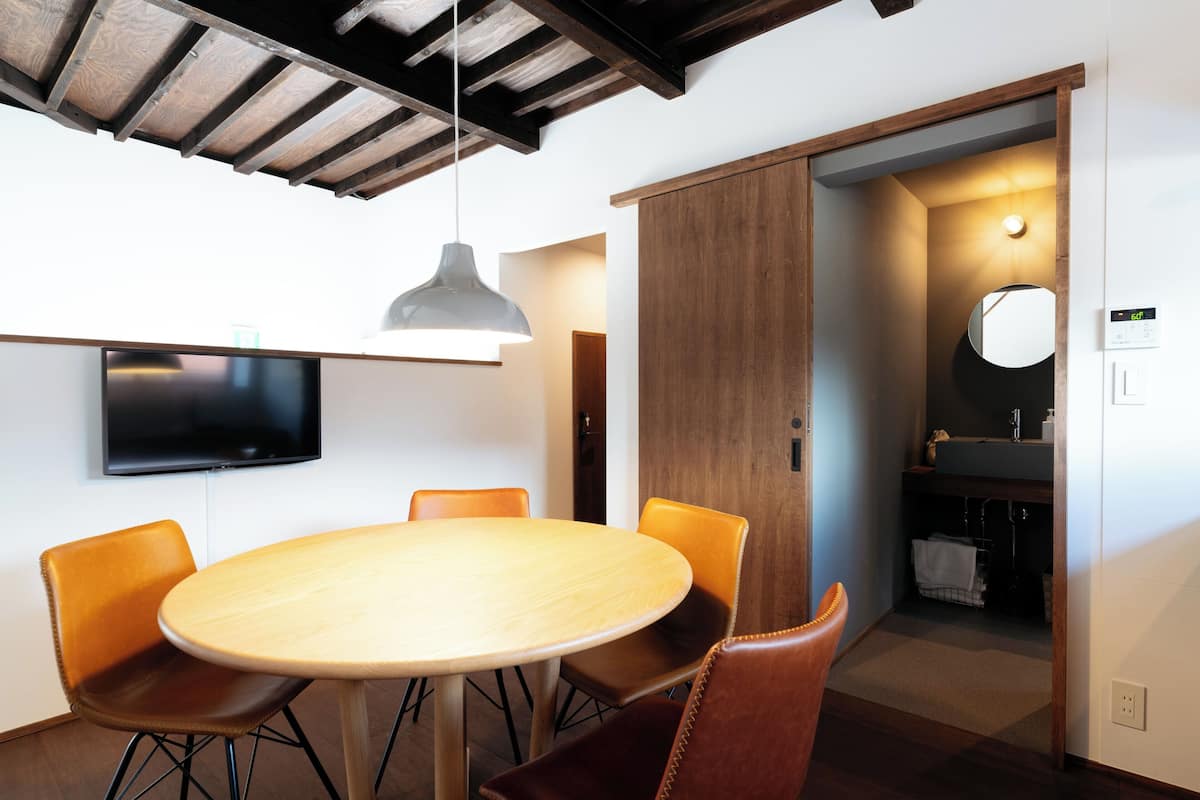 A modern dining area with a round table, leather chairs, a mounted TV, and a wooden ceiling.