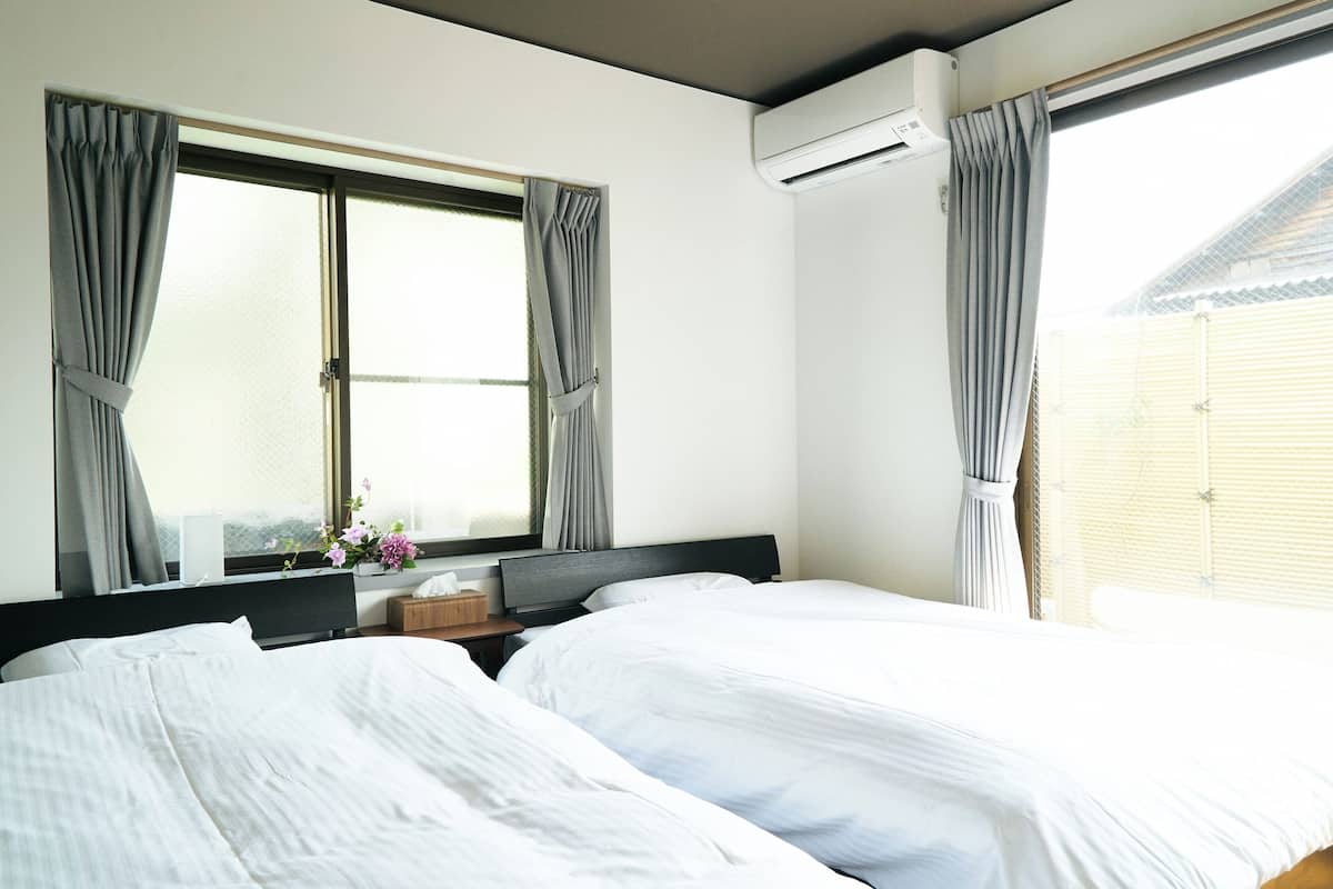 A hotel room with two beds, a window with curtains, an air conditioning unit, and a small table with a vase of flowers.