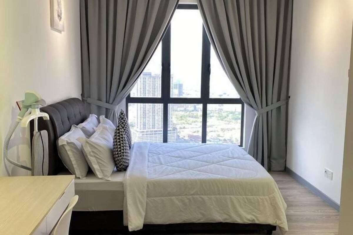 Ein Schlafzimmer mit Bett, Vorhängen, einem Nachttisch und Blick auf Gebäude durch große Fenster.