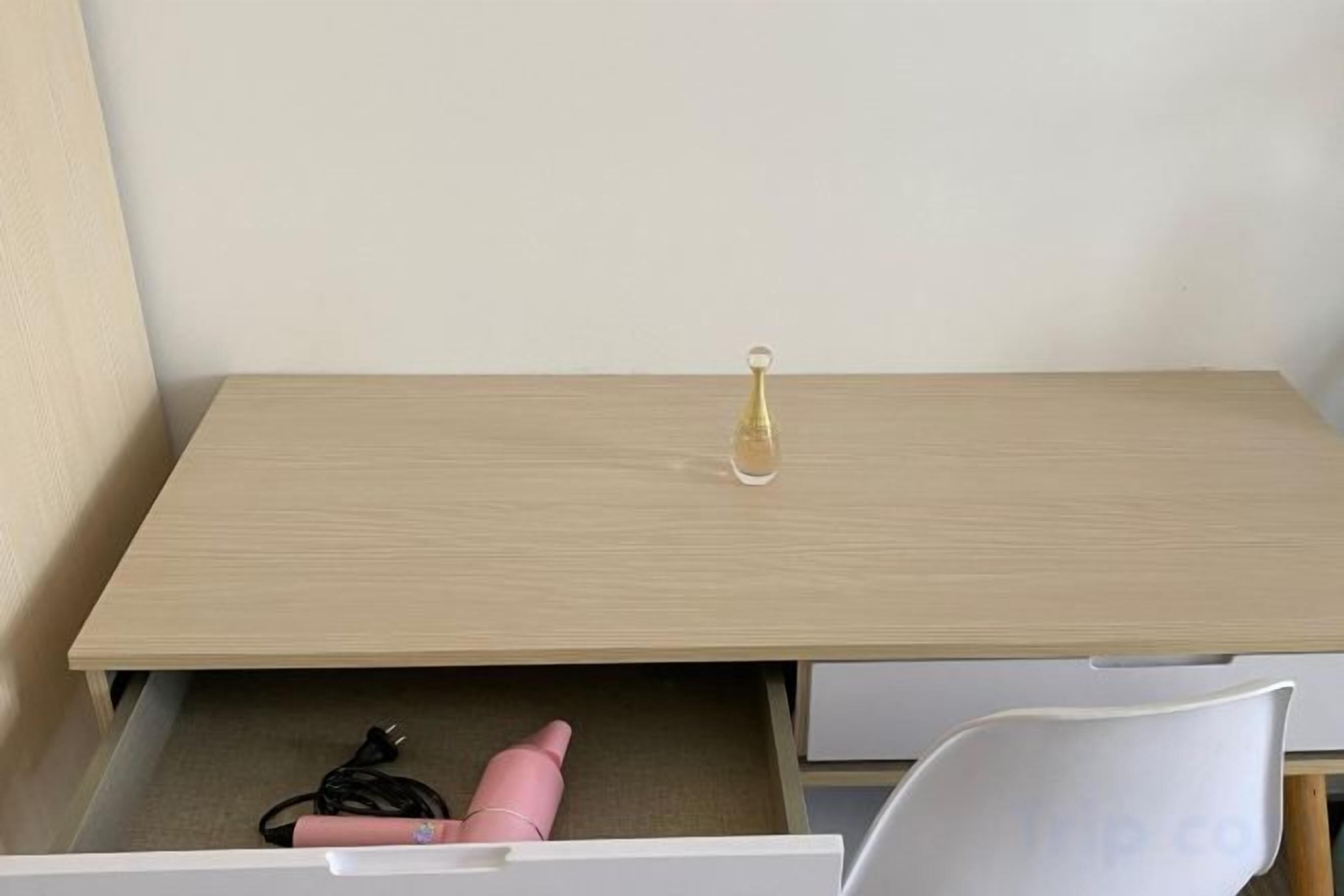 Un escritorio de madera con un cajón, una silla blanca y una pequeña botella sobre la superficie.