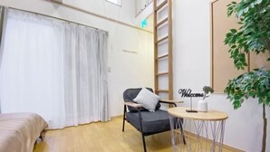 Ein Hotelzimmer mit einem Bett, einem Sessel, einem kleinen Tisch mit einem 'Welcome'-Schild, einer Topfpflanze und einer an der Wand stehenden Leiter.