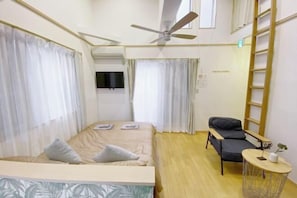 A compact hotel room with a bed, a desk, a chair, a small table, a ceiling fan, and a window.