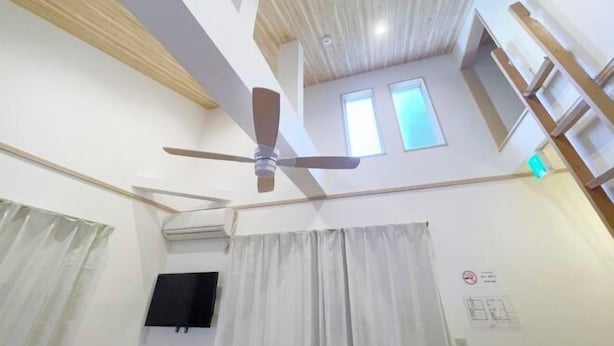 Ein Zimmer mit Deckenventilator, weißen Wänden, einem angebrachten Fernseher und einem Fenster mit Vorhängen.