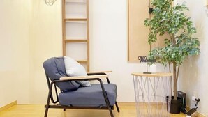 Ein Zimmer mit grauem Sessel, einem kleinen runden Tisch, einem Blumentopf und einem an der Wand befestigten Regal.