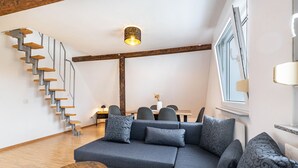 Uma sala de estar moderna com escada de madeira, sofá em formato de L e uma área de jantar ao fundo.