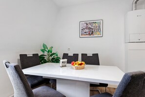 A dining area with a white table, grey upholstered chairs, a plant, and a fruit basket.
