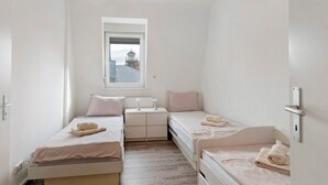Quarto pequeno e branco, com duas camas, uma janela e vista para um edifício.