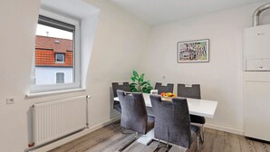 Uma área de refeições com mesa branca, cadeiras cinzas, um radiador e uma janela com vista para um edifício de telhado vermelho.