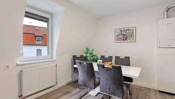 Ein Essbereich mit einem weißen Tisch, grauen Stühlen, einer Heizung und einem Fenster mit Blick auf ein Gebäude mit rotem Dach.
