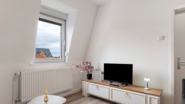 Quarto com radiador branco, armário de madeira com TV, mesa pequena e janela com vista para edifícios.