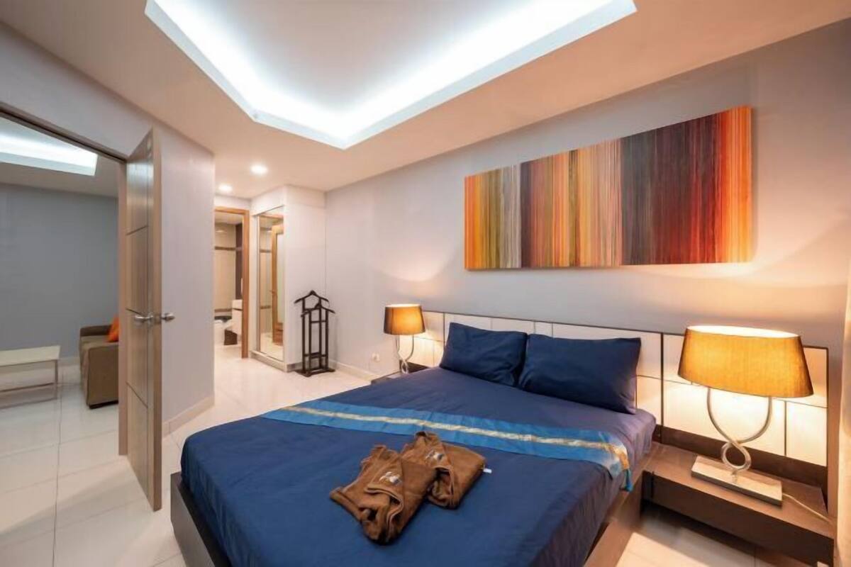 A modern hotel room with a skylight, a large bed with blue bedding, a bedside table with a lamp, and a colorful abstract painting on the wall.