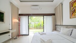 A hotel room with a bed, a TV, a desk, a lamp, and a view of a garden. - Puri Gading Villa Jimbaran by The Lavana (Bali)