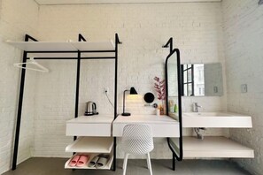 Une pièce de taille modeste, comprenant un bureau blanc, une chaise, un portant à vêtements, un miroir et un lavabo.