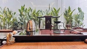 Een tafel met een theepot, een glas en een houten dienblad, met grote groene planten op de achtergrond.
