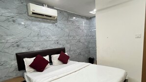 Une chambre avec un mur décoré d’un motif marbré, un lit avec une literie blanche et deux oreillers rouges, une table de chevet et un appareil de climatisation.