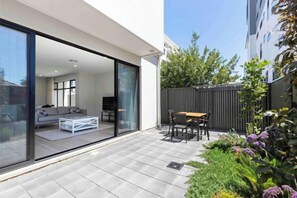 Eine moderne Terrasse mit Glastür zu einem Wohnbereich, einem kleinen Tisch im Freien und einem Garten mit verschiedenen Pflanzen.