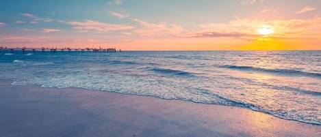 A beach with a pier extending into the water, waves gently lapping at the shore, and a sunset in the background.