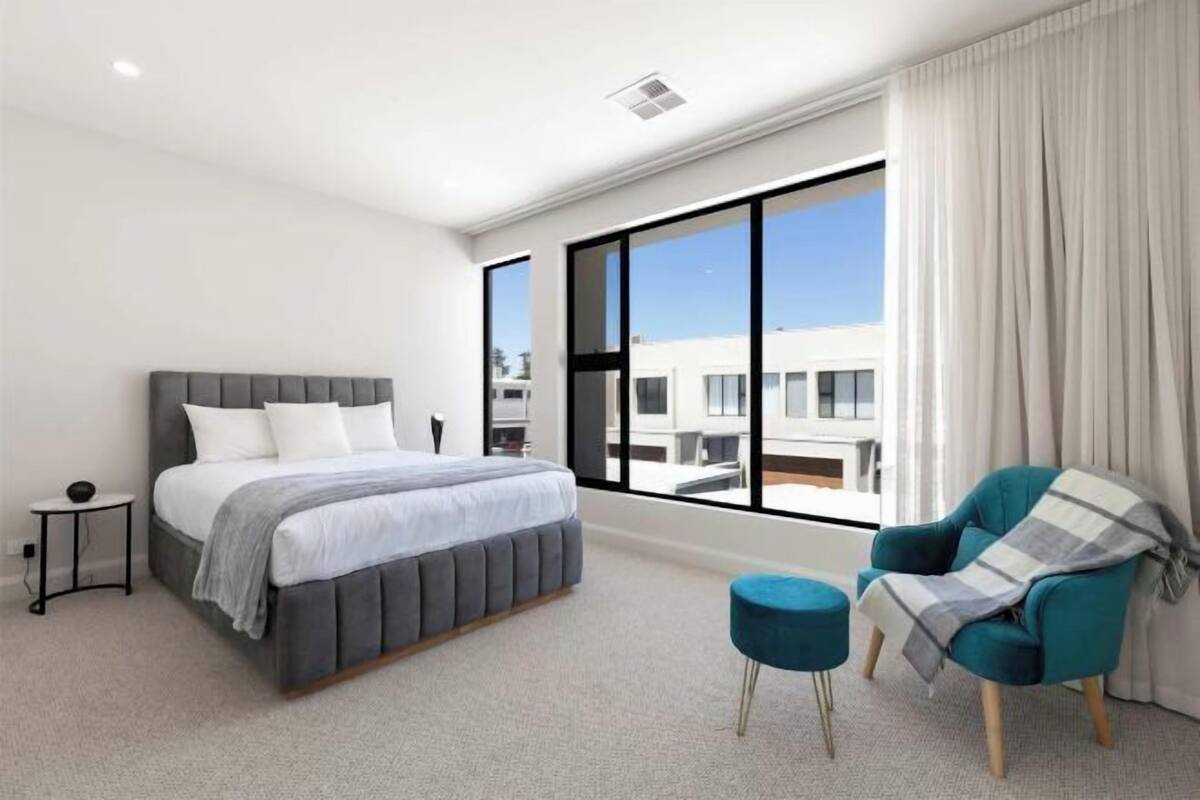 Een moderne slaapkamer met een groot bed, een klein bijzettafeltje, een blauwe poef, een blauwgroene fauteuil en een groot raam met uitzicht op gebouwen.