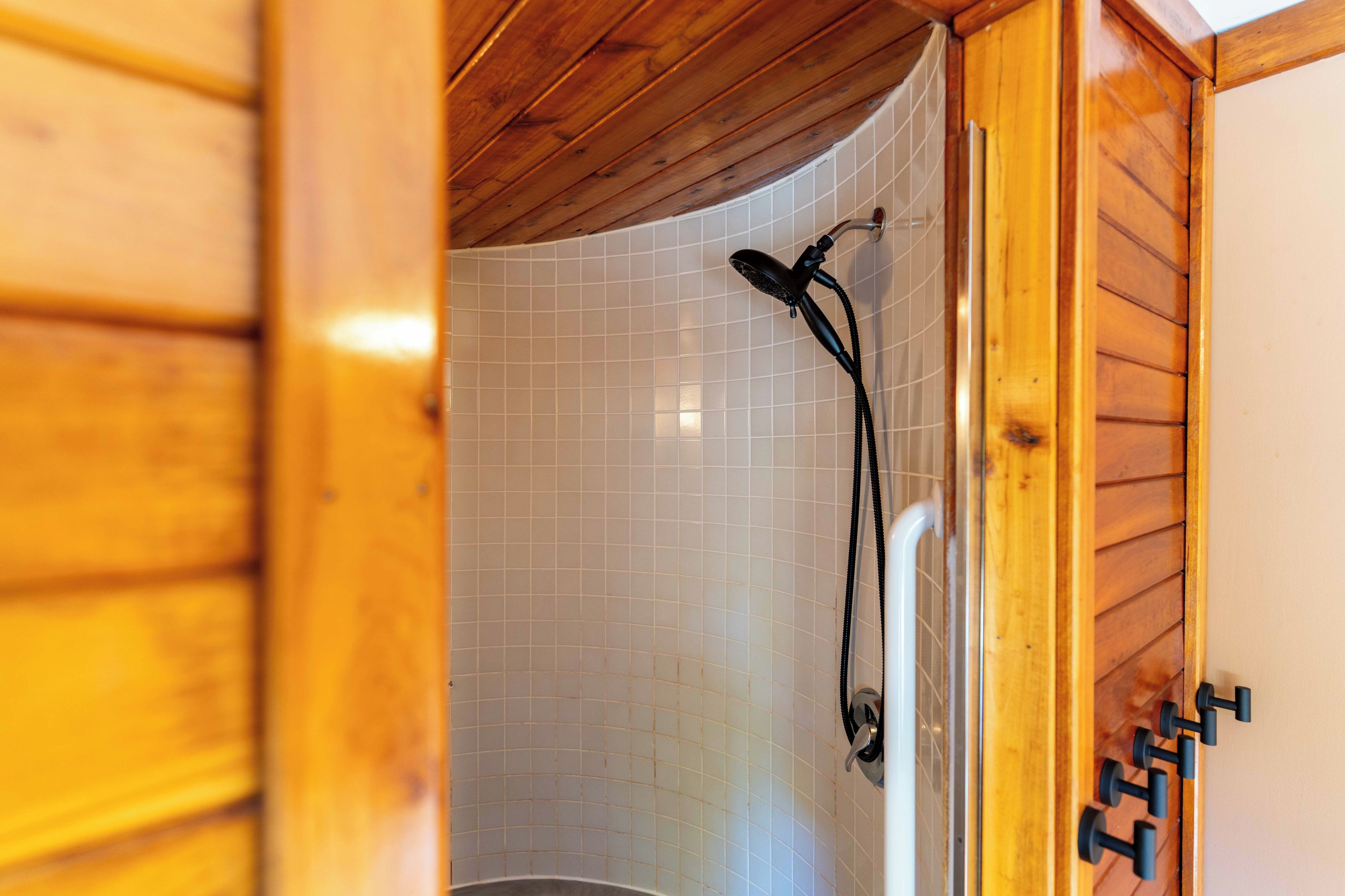 A wooden cabin interior with a shower area featuring white tiled walls and a black handheld showerhead.