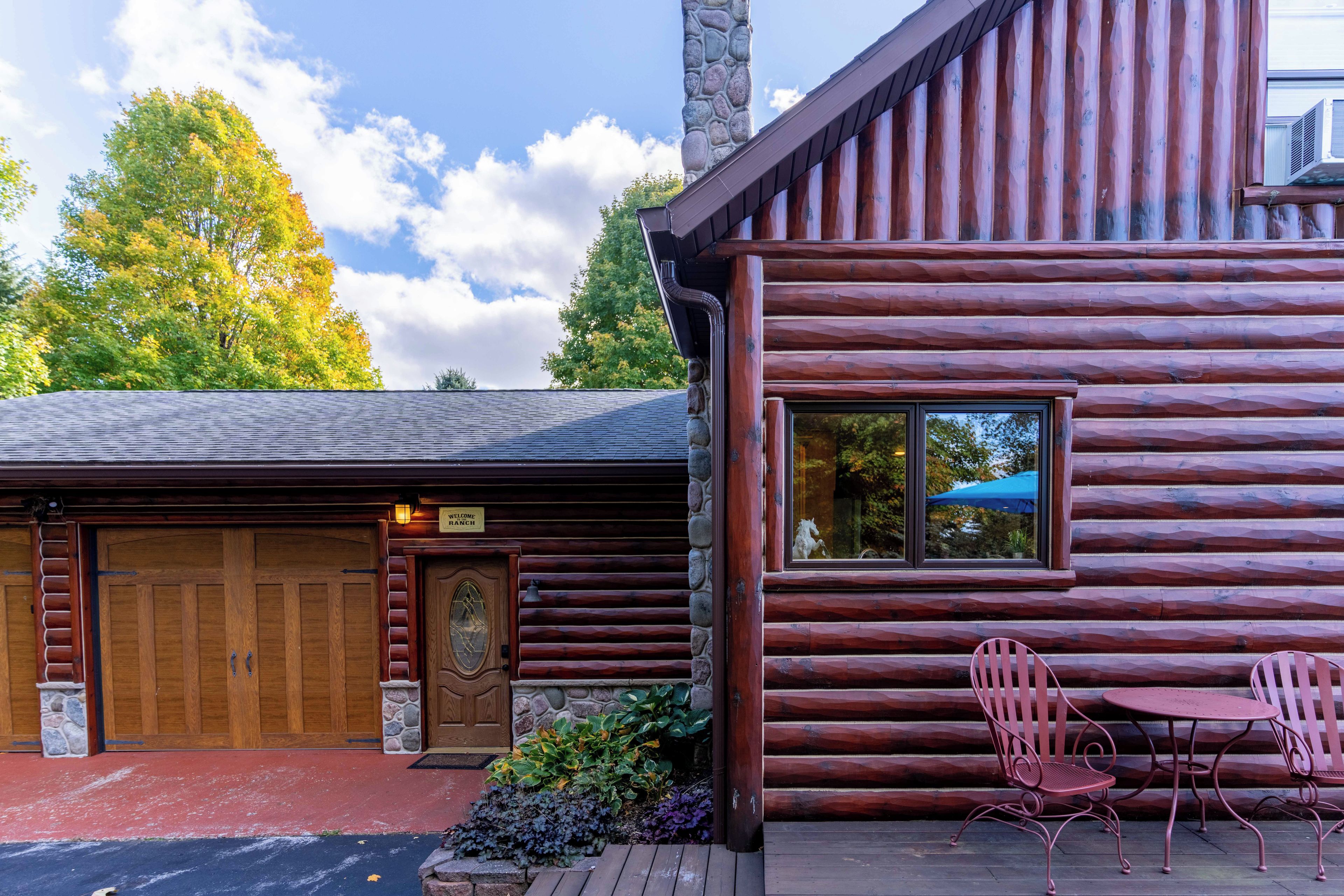 A log cabin with a wooden door, a stone chimney, and a porch with two chairs and a table.