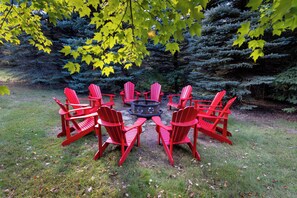 A circular arrangement of red Adirondack chairs around a central fire pit in a grassy outdoor area with trees in the background.