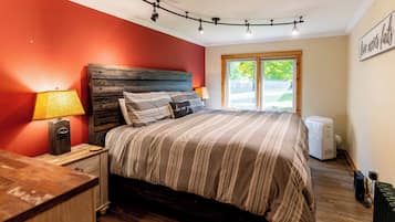 A bedroom with a wooden headboard, a bed with striped bedding, a nightstand with a lamp, a radiator, and a window with a view of greenery.