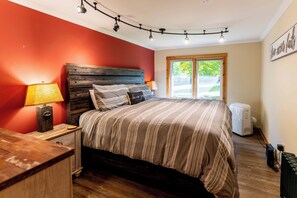 Une chambre à coucher avec une tête de lit en bois, un lit avec une literie à rayures, une table de chevet avec une lampe, un radiateur et une fenêtre donnant sur un paysage verdoyant.