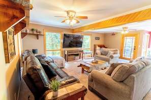 A spacious living room with a large leather sofa, a smaller grey sofa, and a wooden coffee table. A flat-screen TV is mounted on the wall, and there are two windows providing natural light. - NEW! TC Ranch Therapy - Hot Tub, Sauna, Pool Table (Traverse City)