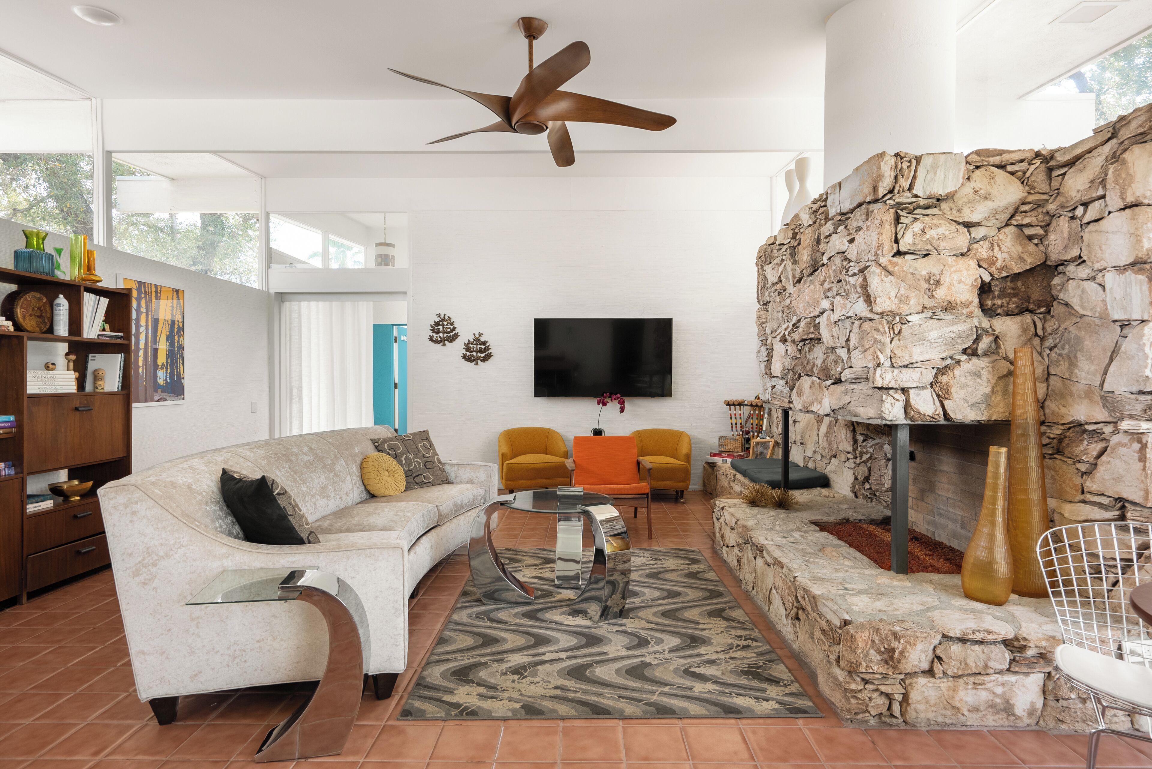 Un salon moderne avec une cheminée en pierre, un canapé blanc, des fauteuils orange et un téléviseur à écran plat fixé au mur.
