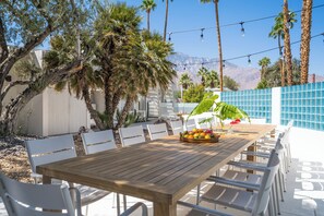 Un espacio de comedor al aire libre con mesa de madera y sillas blancas, rodeado de palmeras y montañas al fondo.