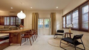 A modern living room with a wooden dining table, chairs, a sofa, and a large window with blinds. - AIRI Home,3BR near Thapae and walking str old town CNX (Chiang Mai)