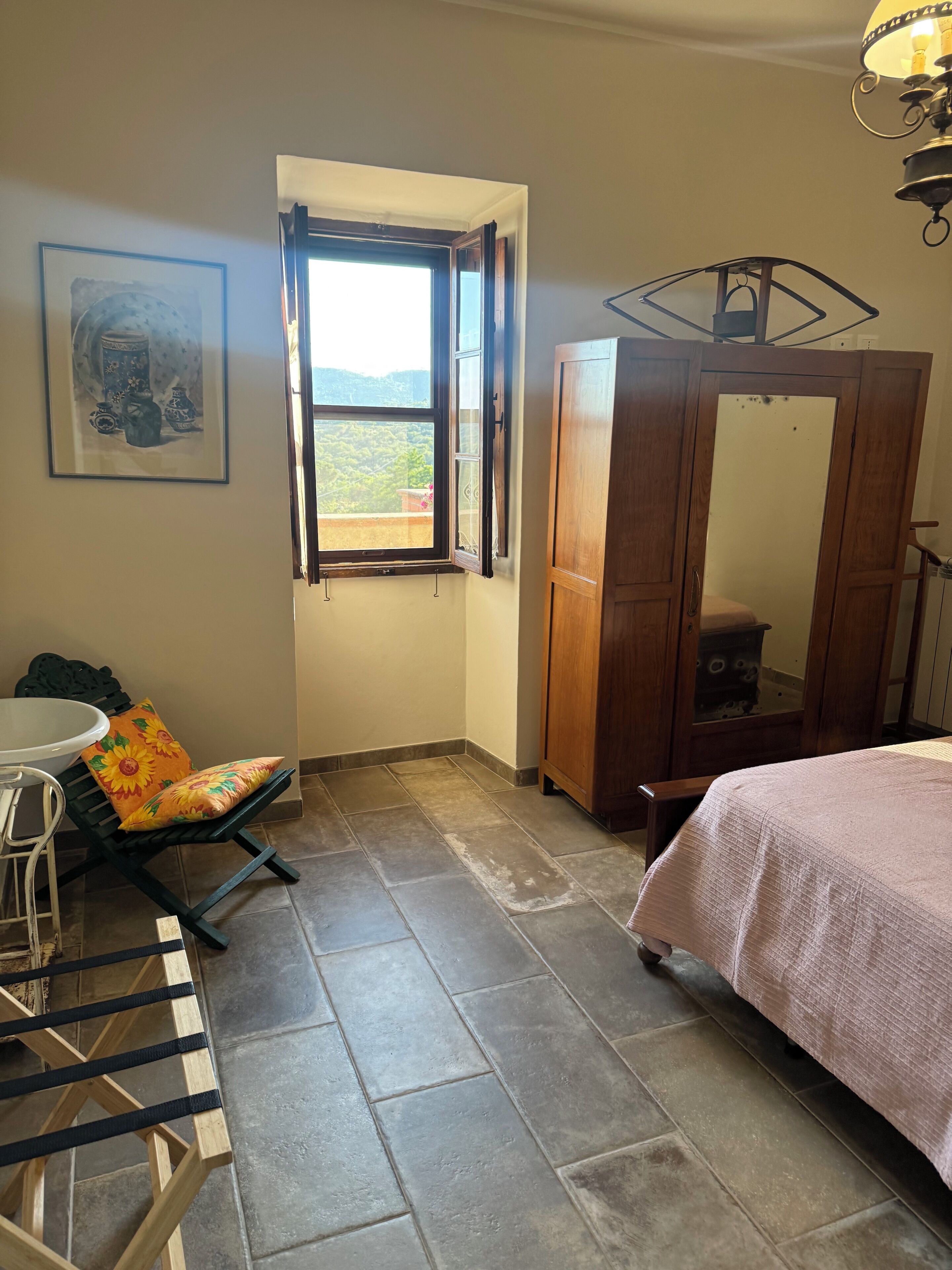 Un dormitorio con cama, armario de madera, silla con cojín, una mesita y ventana con vistas al verdor.