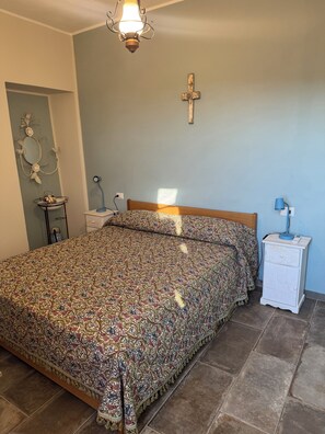 A bedroom with a patterned bedspread, a wooden headboard, a bedside table with a lamp, a wall-mounted cross, and a decorative mirror.
