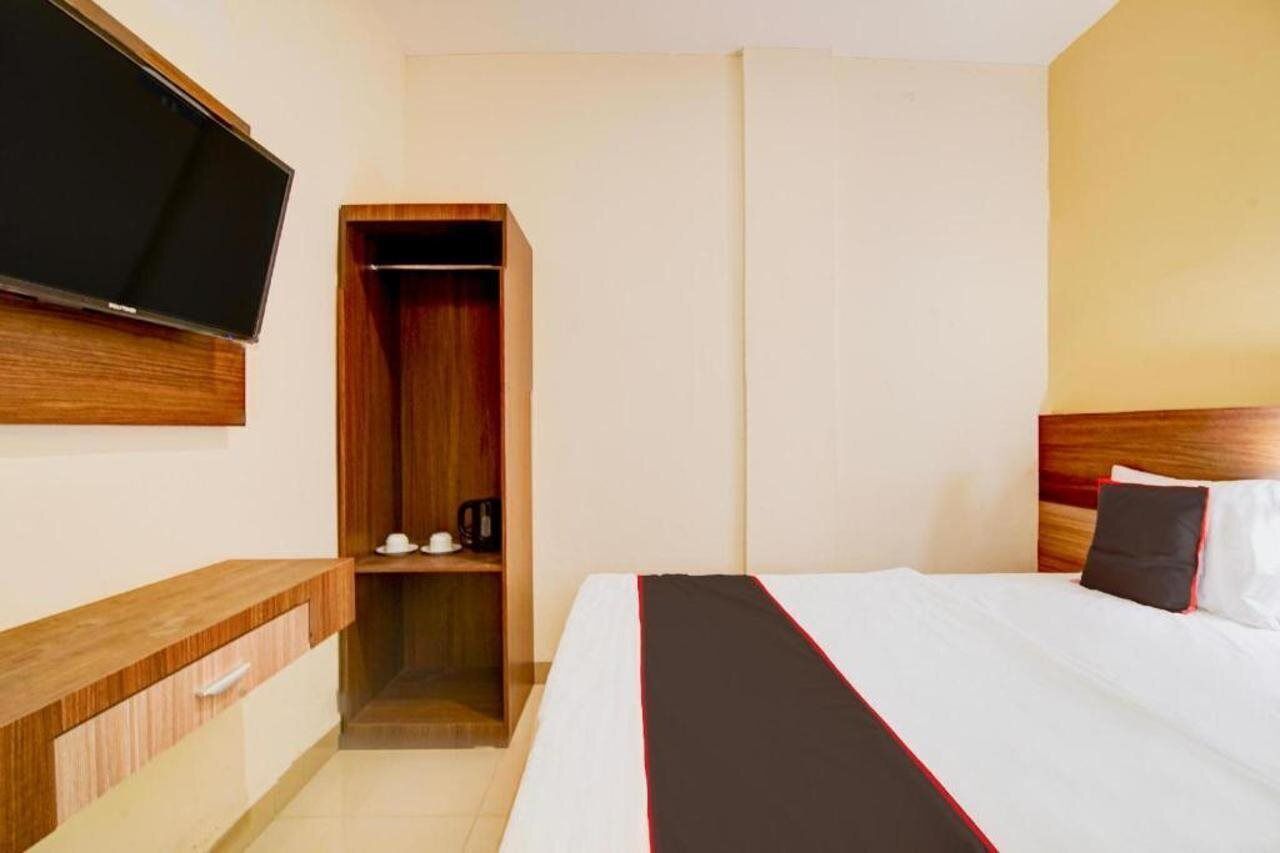 A hotel room with a bed, a TV mounted on the wall, a wooden wardrobe, and a bedside table.