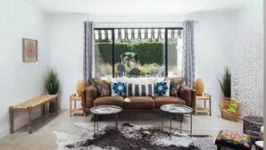 Ein gemütliches Wohnzimmer mit einem braunen Sofa, zwei Beistelltischen, einem Kuhfellteppich und Blick durch ein großes Fenster.